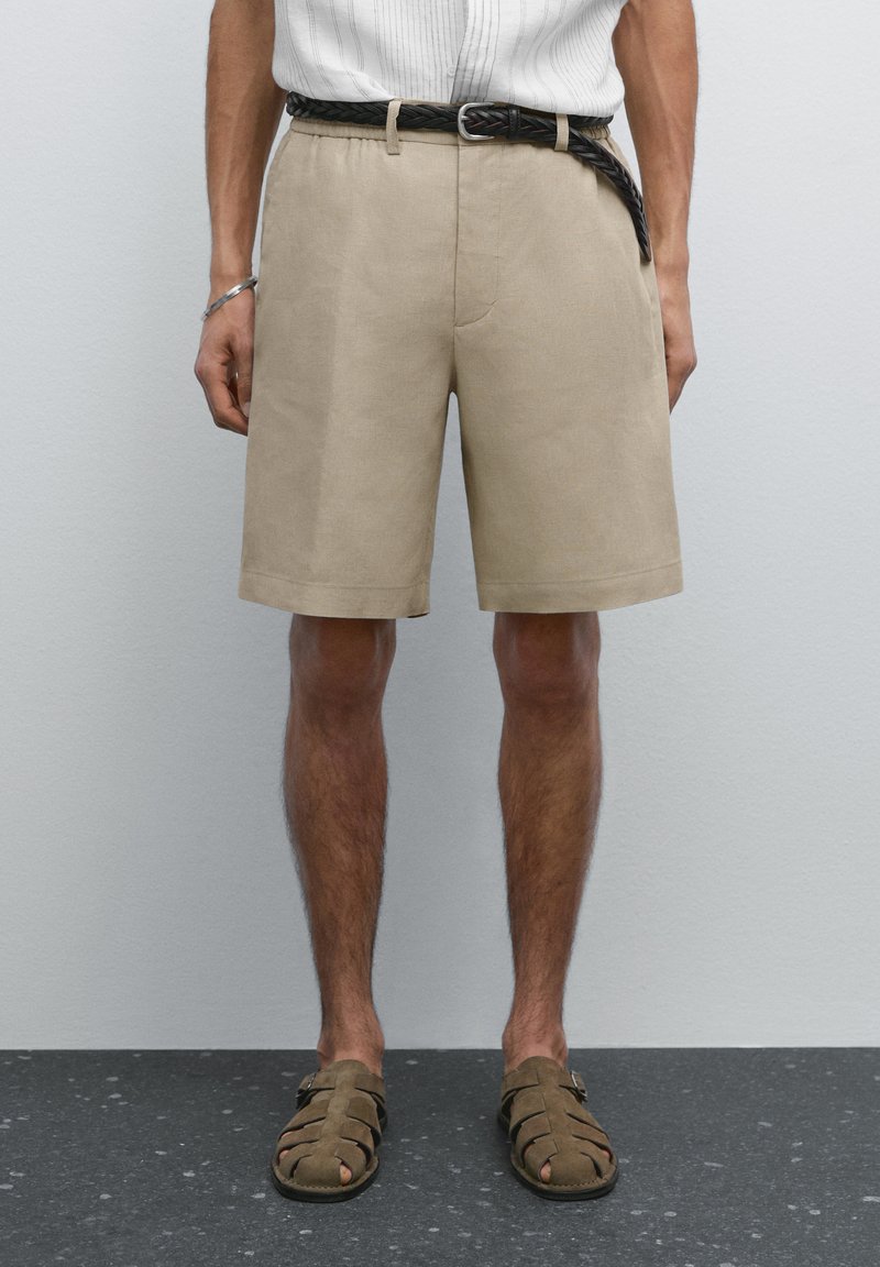 Pantaloni corti beige fino al ginocchio realizzati in tessuto testurizzato, con una vita elasticizzata e una cintura nera intrecciata. Indossati con sandali marroni.