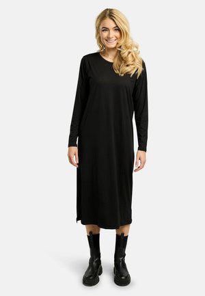 Smilende kvinde med langt blondt hår iført en sort midi-kjole med lange ærmer og sorte chunky ankelstøvler på en hvid baggrund.