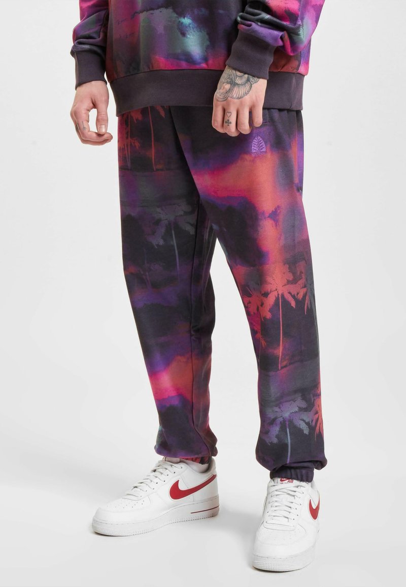 Lilla og svarte joggebukser har et gradientdesign med silhuetter av palmer. Kombinert med hvite Nike-sko med røde detaljer.