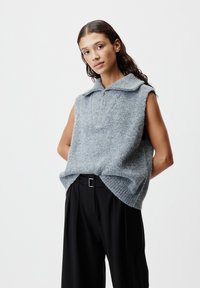Junge Frau in einem lockeren, ärmellosen grauen Strickpullover mit Reißverschlusskragen und schwarzen, weit geschnittenen Hosen, die vor einem schlichten Hintergrund posiert.