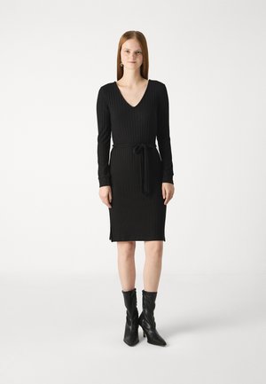 Black ribbed dress with long sleeves, V-neckline, tie waist belt, and side slits. Worn with pointed black leather ankle boots.
