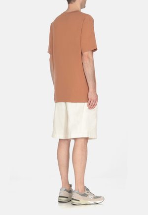 Brązowa bawełniana koszulka o luźnym kroju, zestawiona z białymi szortami i szarymi sportowymi butami. Prosty design, bez widocznych grafik ani wzorów.