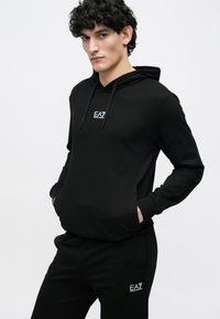Sudadera negra con capucha y cordón, bolsillo delantero y logo en el pecho; combinada con pantalones negros a juego que tienen un logo similar. Tejido suave y elástico.