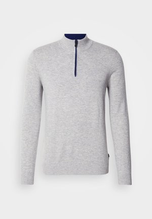 Lichtgrijze, langemouwen trui met een hoge kraag en een rits tot de helft. Marineblauwe accenten bij de kraag. Zachte, gestructureerde stof.