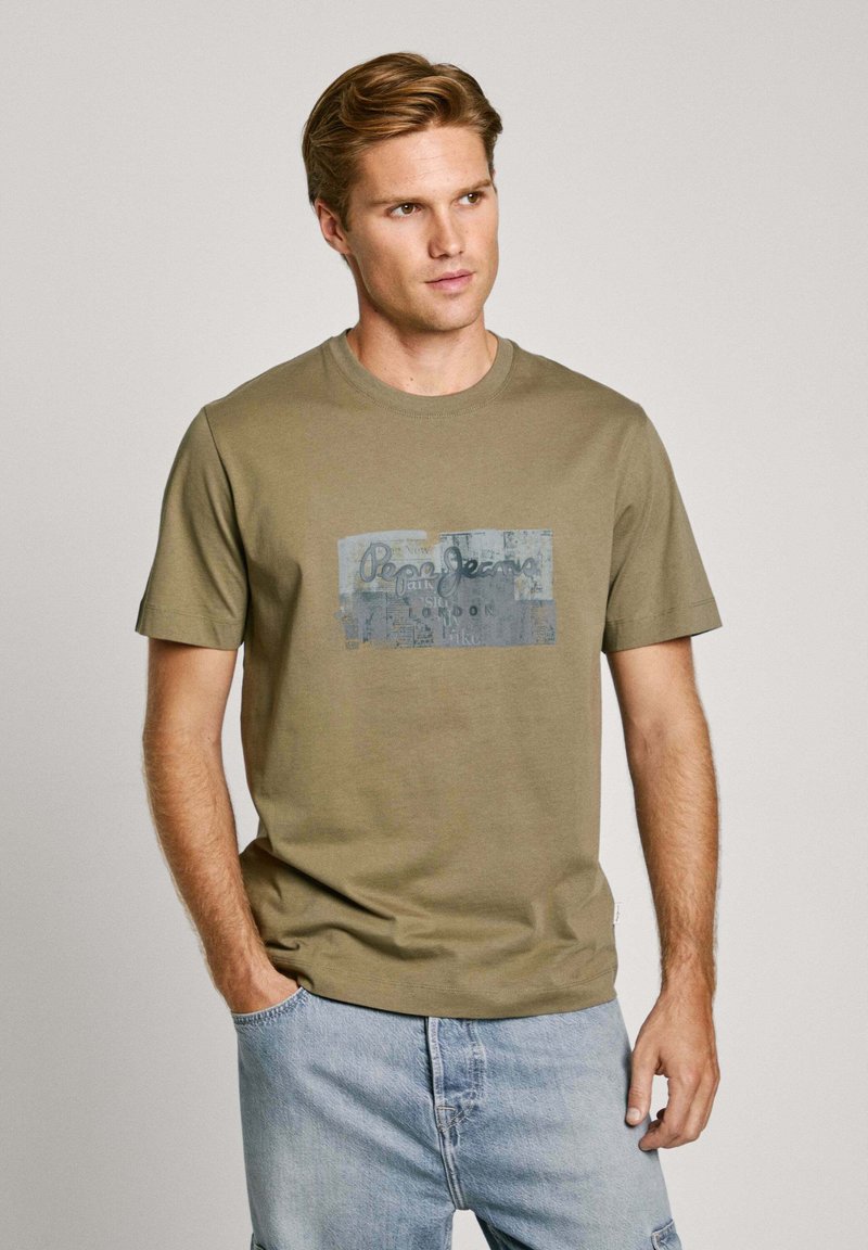 Olijf groene katoenen t-shirt met ronde hals en korte mouwen. De voorkant heeft een bedrukte logo in blauw en grijs met tekst en patchaccenten.