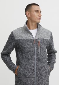 Graue Zip-Jacke mit strukturiertem Design; verfügt über einen hohen Kragen, Seitentaschen und einen kontrastierenden oberen Abschnitt in hellgrau.
