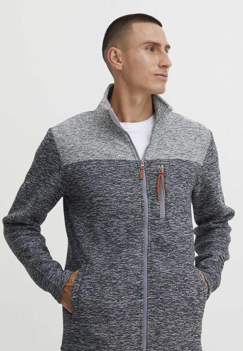 Graue Zip-Jacke mit strukturiertem Design; verfügt über einen hohen Kragen, Seitentaschen und einen kontrastierenden oberen Abschnitt in hellgrau.