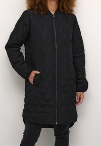 Person trägt eine lange schwarze Steppjacke mit Reißverschluss vorne, Hand in der Tasche, steht vor einer schlichten hellen Wand.