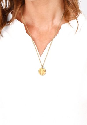 Femme portant un collier chaîne en or avec un pendentif rond en or martelé et un petit charme blanc en forme de goutte, assorti à un chemisier blanc.