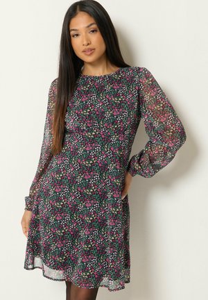Vrouw met lang donker haar die een lange mouwen bloemenjurk draagt met roze, groene en witte bloemen op een lichte achtergrond.