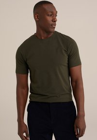 Olivově zelené tričko s krátkým rukávem vyrobené z jemné tkaniny, s klasickým kulatým výstřihem a přiléhavým střihem. Nosí se s tmavými kalhotami.