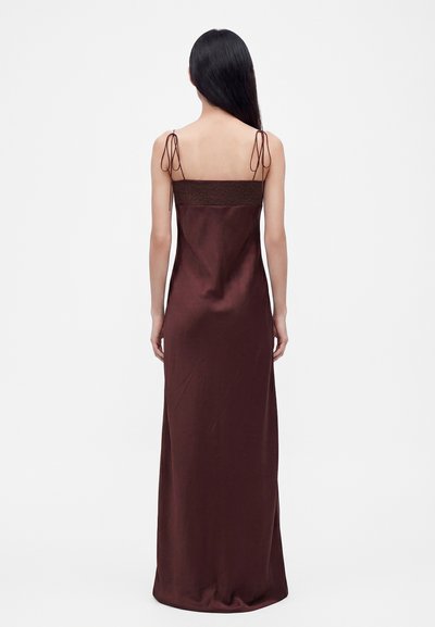 Femme aux longs cheveux noirs portant une robe marron sans manches longue jusqu'au sol avec de fines bretelles nouées aux épaules, de dos sur un fond blanc uni.