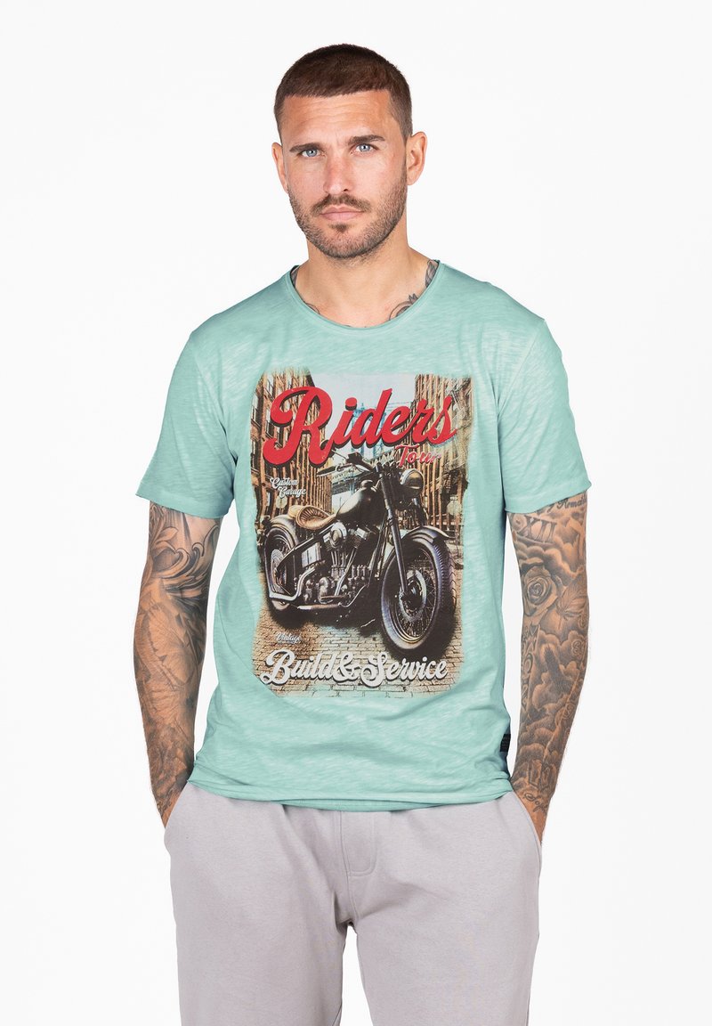 T-shirt verde menta a maniche corte con grafica di una moto vintage, testo "Riders" e "Build & Service" in rosso e bianco. Texture morbida.