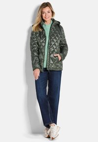 Grüne gesteppte Jacke mit Kapuze, Frontreißverschluss und zwei Seitentaschen. Getragen über einem hellgrünen Pullover, kombiniert mit dunkelblauen Jeans und Sneakers.