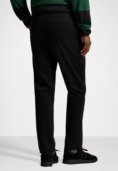 Black trousers with a relaxed fit, featuring back pockets. The fabric appears smooth, with a subtle sheen. Worn with black slip-on shoes.