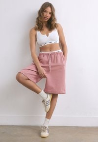 Shorts en coton rose avec une taille élastique, détail logo et poches latérales, associés à un haut court blanc et des baskets de couleur claire.