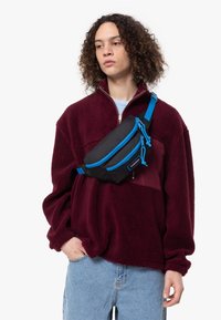 Černá ledvinka s jasně modrými zipy a popruhem, s hladkým povrchem, nošená diagonálně přes burgundskou fleecovou bundu.