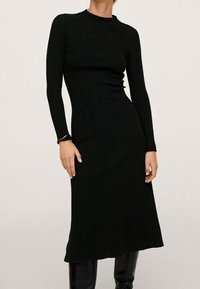 Femme portant une robe midi noire en maille côtelée à manches longues près du corps, associée à des bottes jusqu'aux genoux noires, sur un fond clair uniforme.