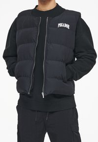 Svart uppblåsbar väst med dragkedja fram, vadderad design och logotyp på vänster bröst. Bärs över en svart långärmad skjorta och svarta byxor.