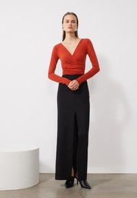 Red long-sleeve top with a wrap design and gathered detail, paired with a high-slit black skirt. The overall look is sleek and contemporary.