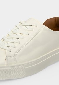 Baskets basses blanches avec lacets blancs, tige en cuir lisse, semelle en caoutchouc texturée et doublure intérieure beige visible au talon.