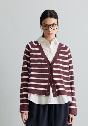 Frau mit dunklen Haaren und Brille, die eine karminrote und weiß gestreifte Strickjacke über einem weißen Hemd mit Knöpfen und dunkler Hose trägt, vor einem grauen Hintergrund.