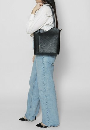 Zwarte, gestructureerde tote bag met een gebogen bovenkant, voorzien van een zipvak en lange schouderriemen, gecombineerd met lichtblauwe jeans.