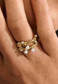 Bague en or présentant un design torsadé, agrémentée de petites perles blanches. Présentée sur une main à la peau texturée, mettant en valeur les détails de la bague.