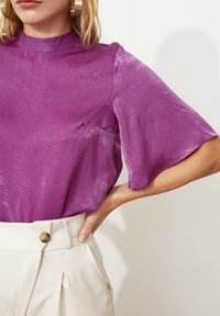 Top in raso viola con maniche corte a campana e scollatura alta; presenta una tasca laterale e si abbina a pantaloni beige a vita alta.