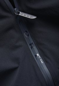 Primer plano de cremallera negra Gore-Tex con pestaña de tirador texturizada sobre tela oscura, mostrando la marca Gore a lo largo de la costura de la cremallera.
