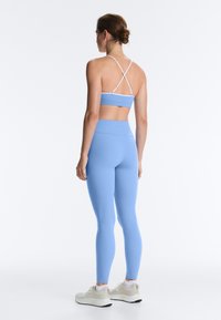 Woman standing, wearing light blue high-waist leggings and a matching sports bra with white crisscross straps, beige sneakers, and white socks.
