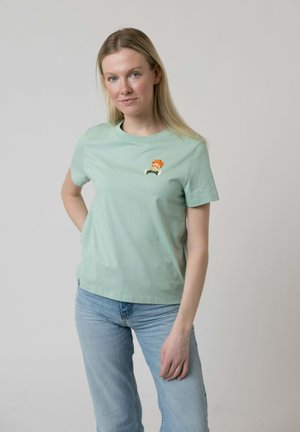 Blonde woman wearing light green t-shirt with small cartoon lion graphic and light blue jeans, standing against plain background.