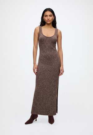Femme portant une robe maxi marron en maille sans manches avec une fente latérale, assortie à des bottines marron, debout devant un fond blanc uni.