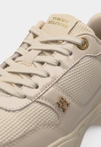 Baskets beiges Tommy Hilfiger avec texture mesh et cuir, lacets blancs, logo doré sur la languette et le côté, et œillets dorés.