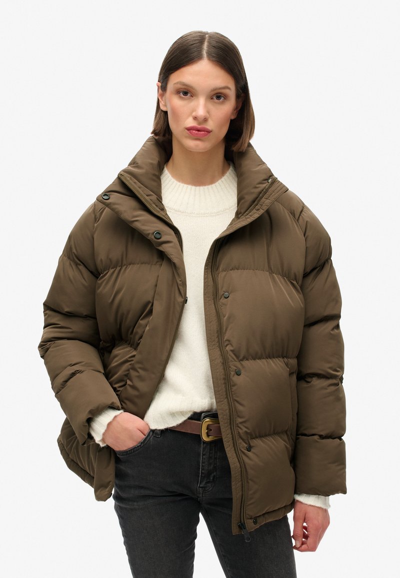 Chaqueta marrón acolchada con un alto cuello, cierres a presión y cremallera. Llevada sobre un suéter ligero. Presenta un corte relajado y una tela texturizada.