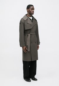 Dark brown trench coat with double-breasted buttons, wide collar, and belted waist. Model stands in relaxed fit black trousers and shoes.