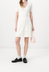 Robe blanche avec design en maille texturée, manches courtes en dentelle et col en V. Portée avec des mocassins noirs et un petit sac à main rose.