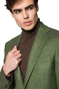 Zielona marynarka z tweedu z wyciętym kołnierzem, zestawiona z brązowym golfem. Zauważalny detal ręczny trzymający za kołnierz marynarki.