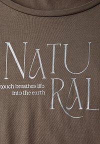 Camiseta marrón con letras grandes en blanco que dicen "NATURAL" con un lema en texto más pequeño: "el toque da vida a la tierra."