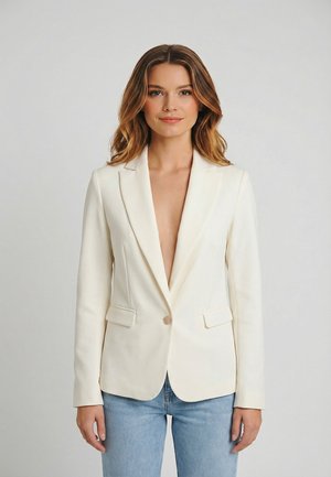 Mujer con cabello ondulado hasta los hombros, vistiendo un blazer crema de un solo botón y jeans azul claro, de pie frente a un fondo claro y sencillo.