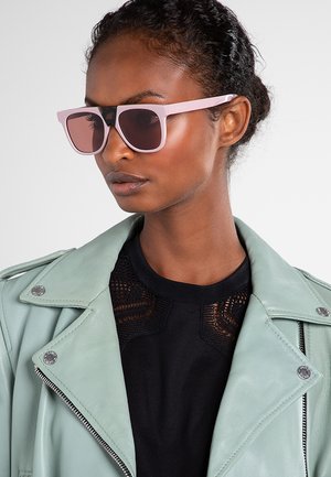 Donna che indossa occhiali da sole oversize rosa chiaro, top in pizzo nero e giacca di pelle verde menta con bottoni e zip in metallo.