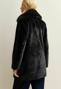 Blondynka w czarnym, puszystym płaszczu z sztucznego futra, stojąca bokiem z ręką swobodnie opuszczoną wzdłuż ciała na tle jasnej, gładkiej ściany.