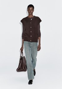 Kobieta w brązowej, zapinanej kamizelce, nosząca długi rękaw bluzki, jasnoniebieskie dżinsy, czarne buty i trzymająca dużą, ciemnobrowną torebkę.