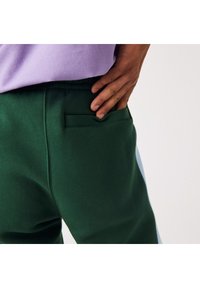 Gröna träningsbyxor med en smal, horisontell ficka; mjuk textur med en slät yta och ljusblå sidopanel.