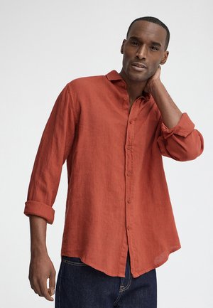 Man draagt een roest-oranje overhemd met opgerolde mouwen en donkerblauwe spijkerbroek, poseert met één hand die zijn nek raakt.