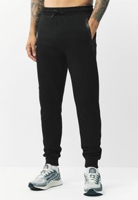 Schwarze Jogginghose mit Kordelzugbund, seitlichen Reißverschlusstaschen und schmal zulaufenden Bündchen. Hergestellt aus einem weichen Stoff mit glatter Textur.