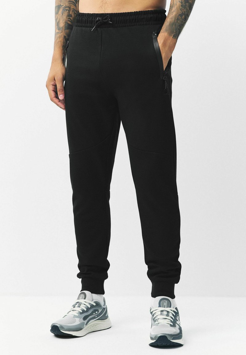 Schwarze Jogginghose mit Kordelzugbund, seitlichen Reißverschlusstaschen und schmal zulaufenden Bündchen. Hergestellt aus einem weichen Stoff mit glatter Textur.
