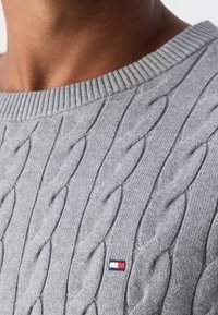 Gros plan d'une personne portant un pull en maille torsadée gris clair avec un petit logo rectangulaire rouge, blanc et bleu sur la poitrine.