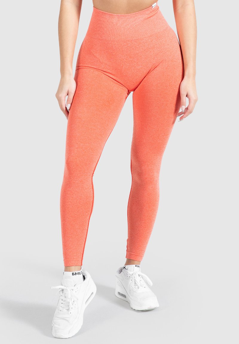 Hoge taille oranje leggings van een zacht, rekbaar materiaal met een gladde textuur en naadloos ontwerp, gecombineerd met witte sportschoenen.