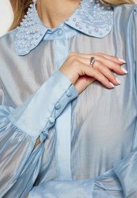 Blusa blu chiaro con colletto decorato di perle, polsini con bottoni e tessuto morbido e liscio con una leggera lucentezza.